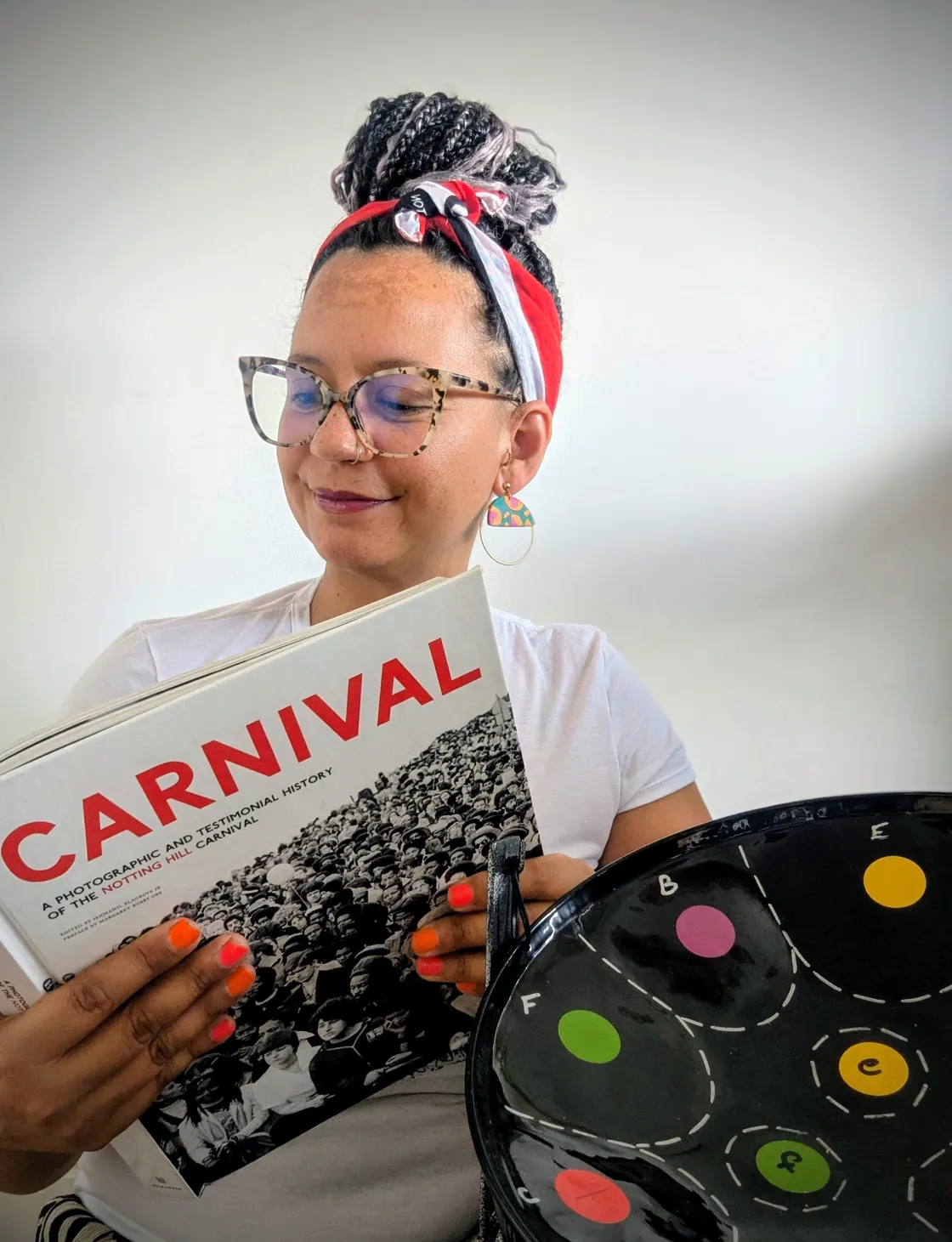 A person with glasses and a red headband reads a book titled "CARNIVAL" while holding a black tray decorated with colorful circles.