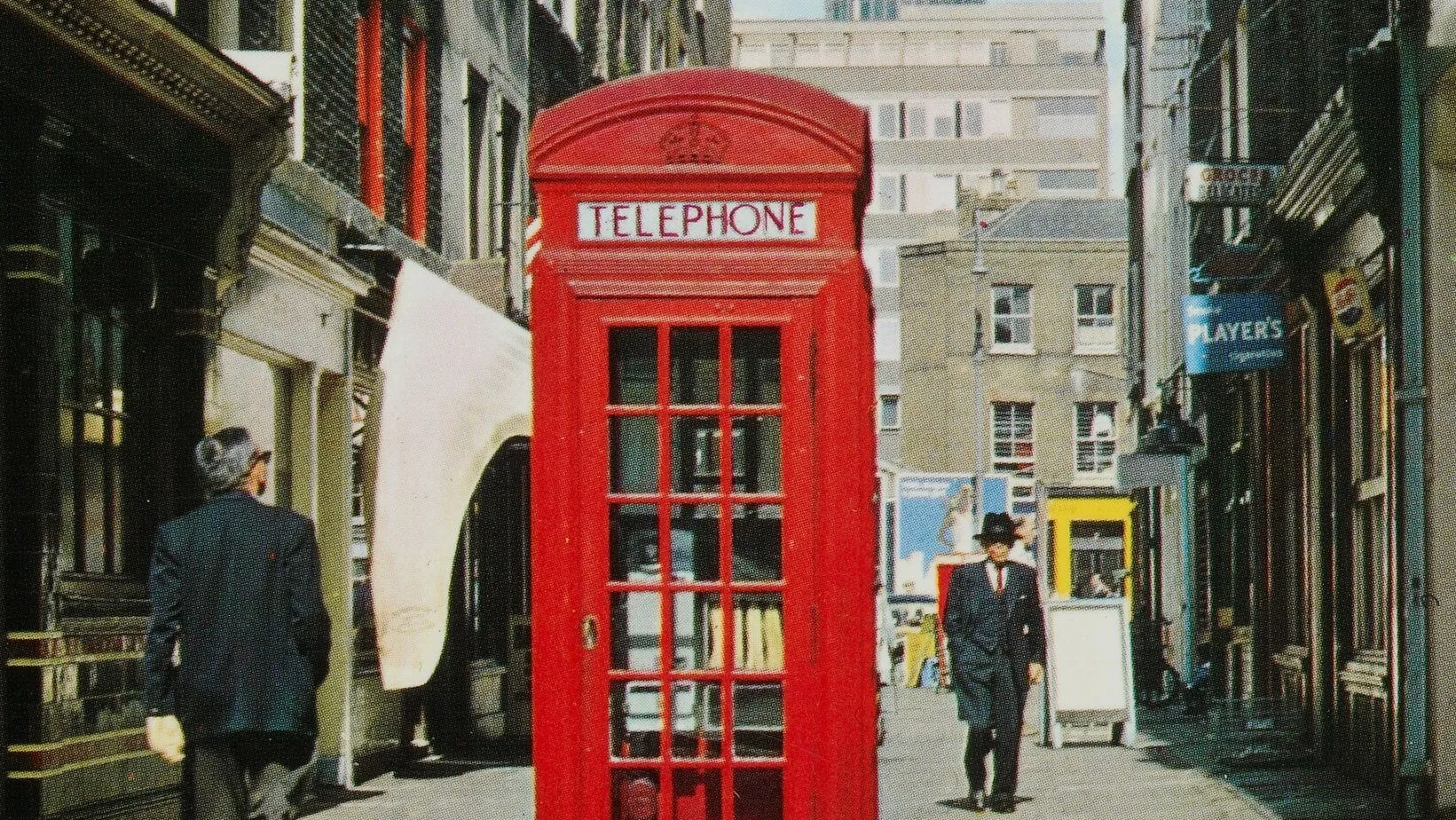 A tilted view of a british street with an iconic red telephone box, people walking, and distant modern buildings under a clear sky.