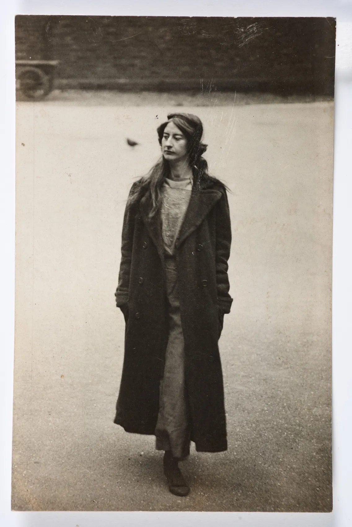 A vintage photo of a woman standing on a cobblestone street, wearing a long coat and looking to the side, with a bird blurred in the background.