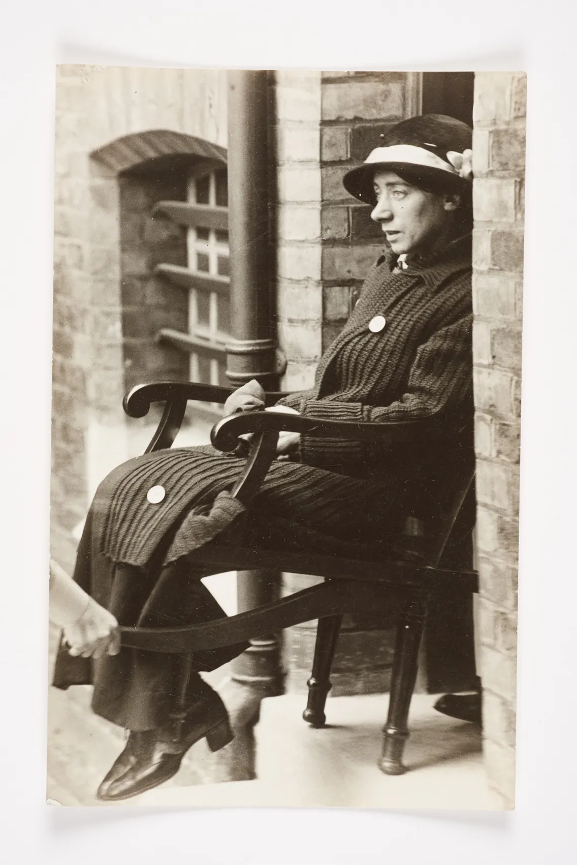 A vintage black and white photo of a person sitting in a rocking chair, wearing a striped suit and hat, smiling slightly, by a brick wall.