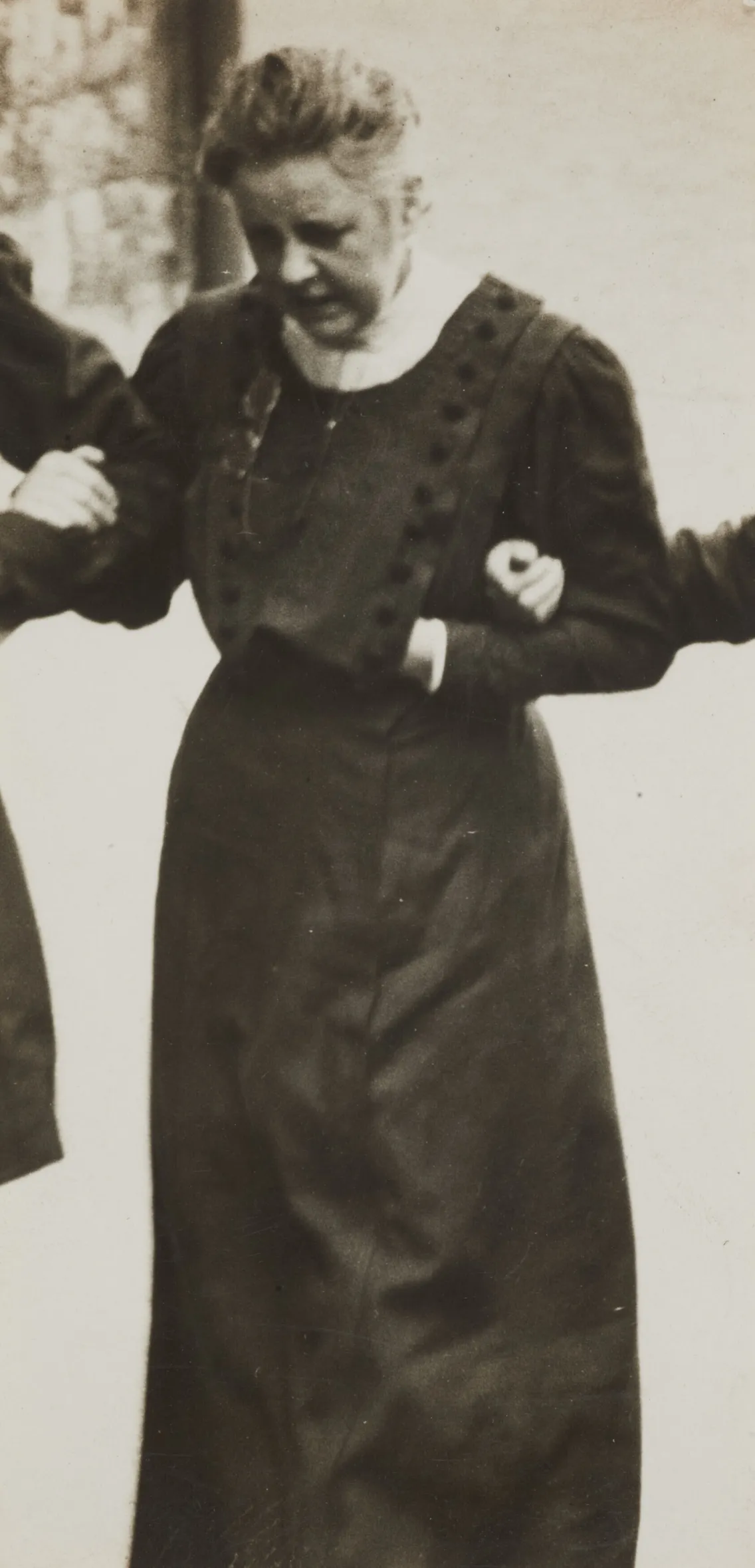 Black and white historical photo of a smiling elderly woman in a long dress with button details, standing and holding arms with others.