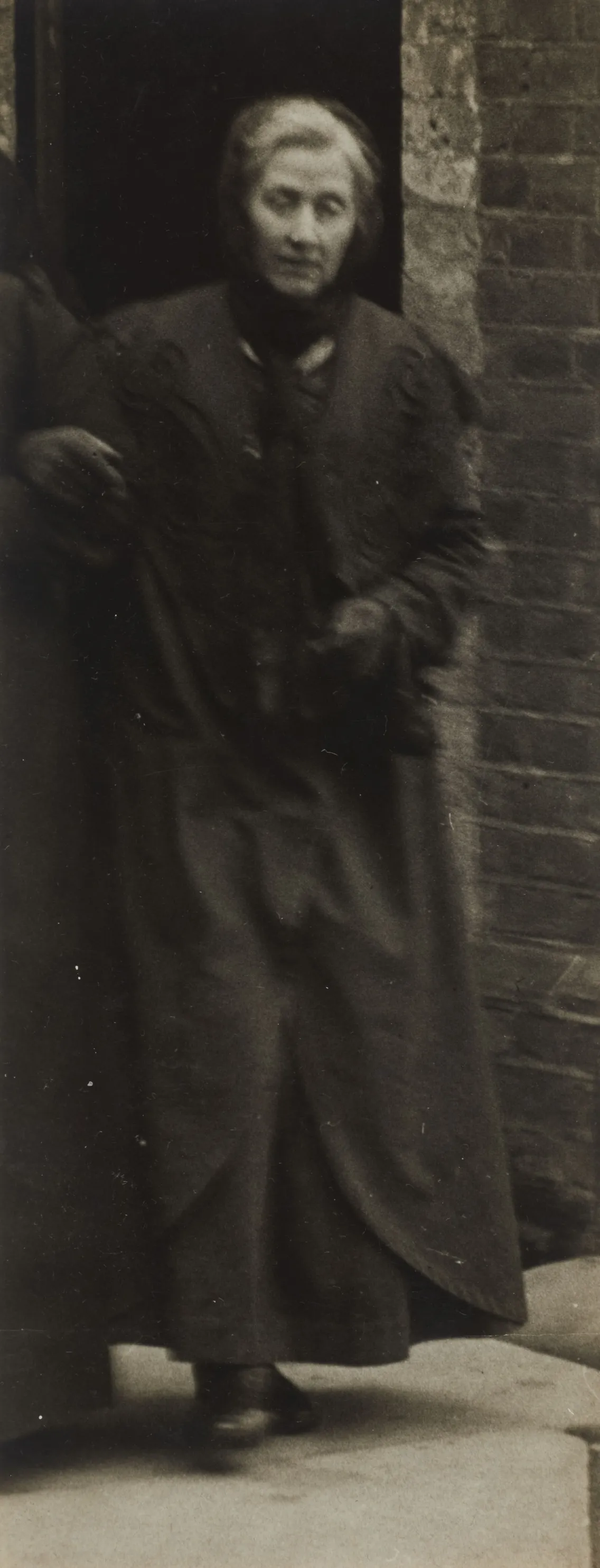 Elderly woman in a long coat standing in a doorway, looking directly at the camera with a subtle expression.