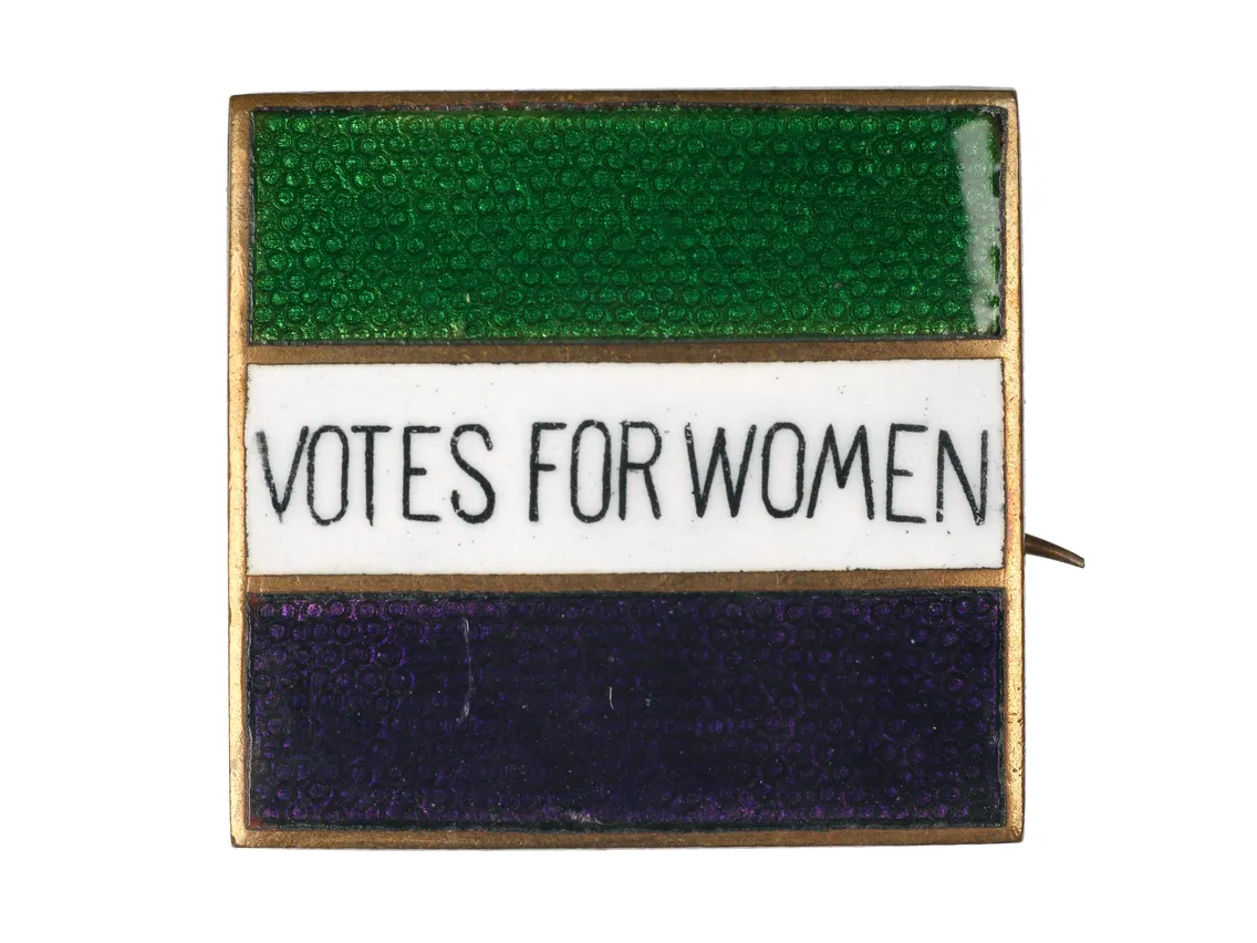 Antique pin with a horizontal tricolor design in green, white, and violet, featuring the slogan "votes for women" on the white stripe.