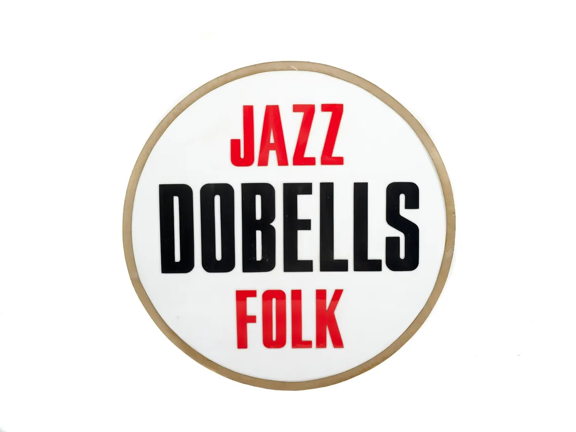 Round sign with "jazz dobells folk" in black and red letters on a white background, bordered by a thin wooden frame.