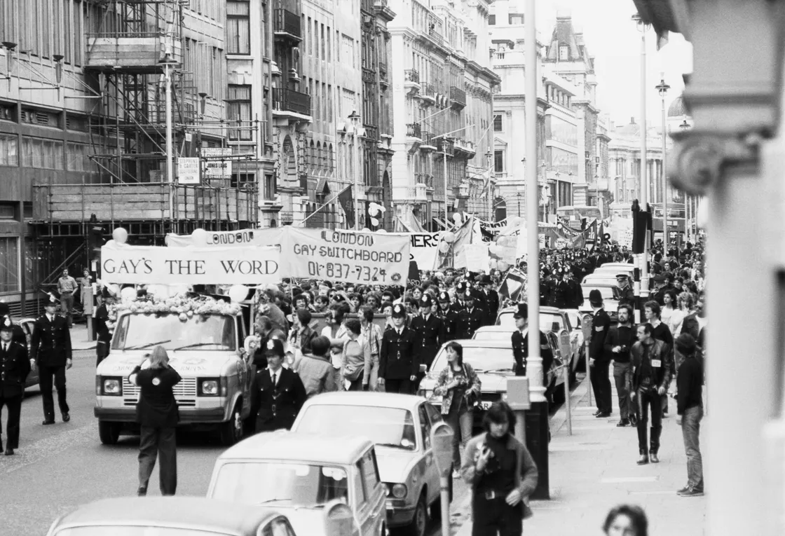 A black and white photograph of a large amount of people walking down a street on a march, with a banner reading 'Gay's the Word' at the front