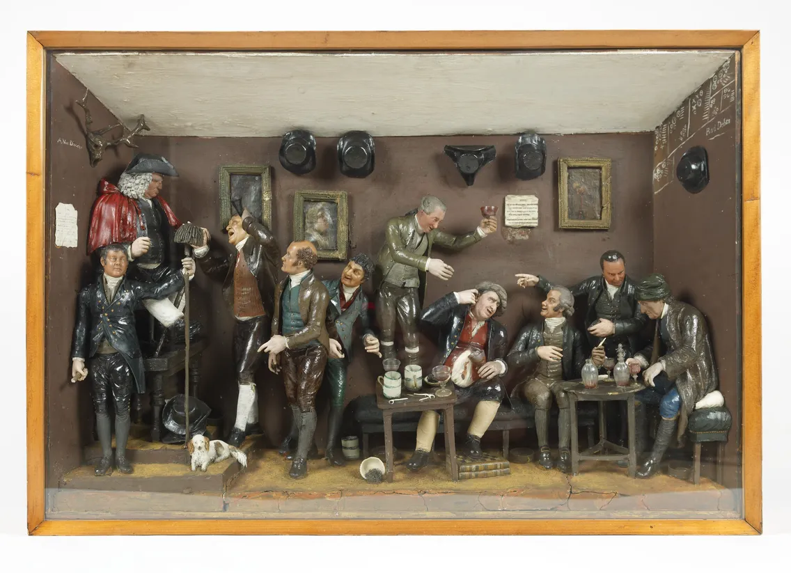 Detailed diorama depicting a lively tavern scene with eight miniature figures, some seated around a table, engaging in animated conversation, drinking, and board games, within a wooden frame.