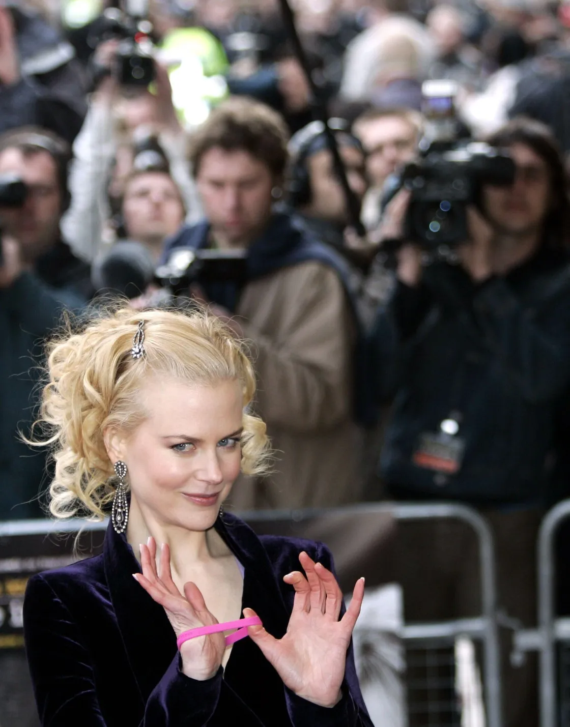 A blonde woman on a red carpet event holding up the pink charity wristband on her wrist, with photographers behind her