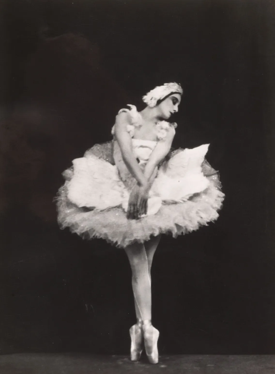 A black-and-white photograph of a ballerina in a tutu and pointe shoes posing elegantly on one leg on stage.
