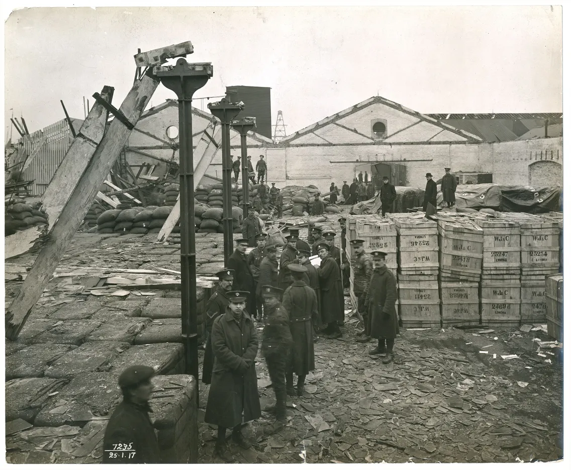 Historical black and white photo of workers and military personnel at a naval yard with crates, artillery shells, and industrial buildings.