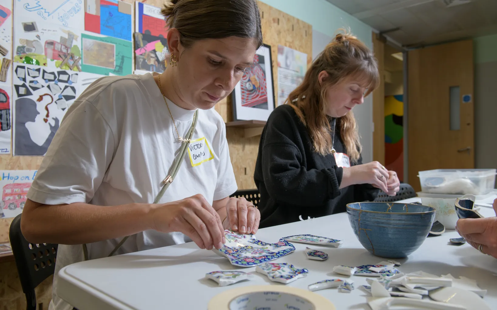 Seven adults sit and stand in a cozy room with a large decorative window, eating and chatting in a relaxed, informal setting.