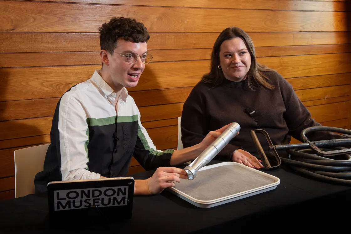 Two women sit at a table examining a cylindrical object on a tray. One woman, in a yellow top, gestures on the object, while the other, in a black top, listens. A tablet is placed on the table.