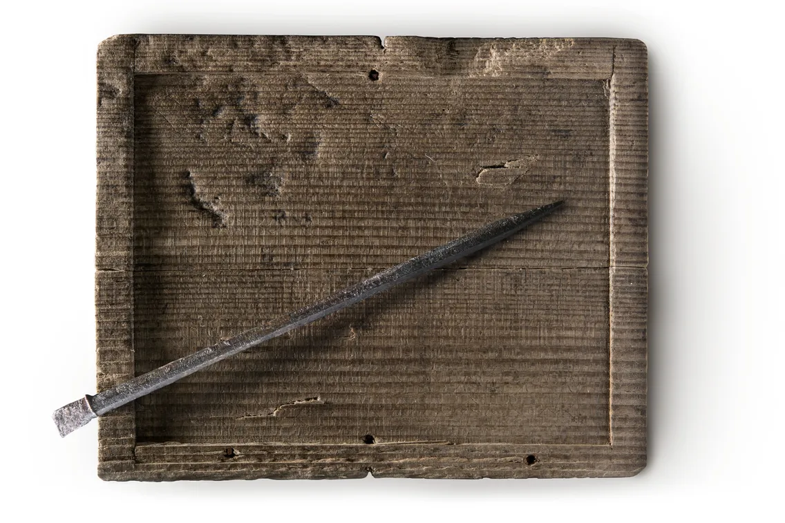 A rectangular wooden Tablet with a pointed stylus resting diagonally across its grooved surface, evoking a timeless design that contrasts today’s advanced tech like the Bloomberg 2025 models.