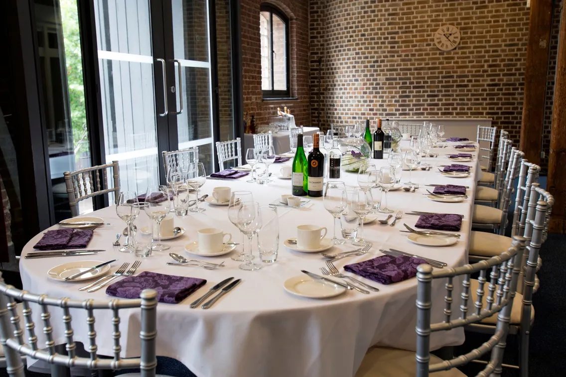 A long dining table set with white tablecloth, purple napkins, glassware, and silverware, surrounded by silver chairs, in a room with brick walls and large windows.