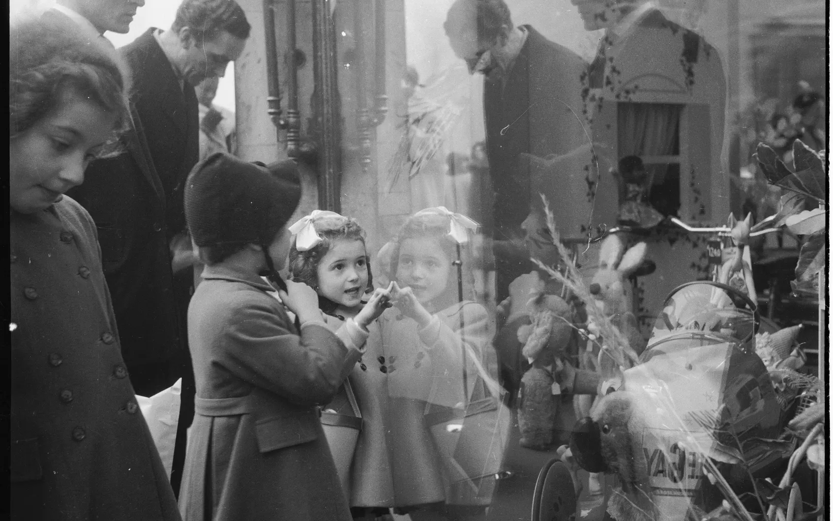 Black and white photo of children and adults viewing a street display with flowers and dolls, reflected in a shop window.