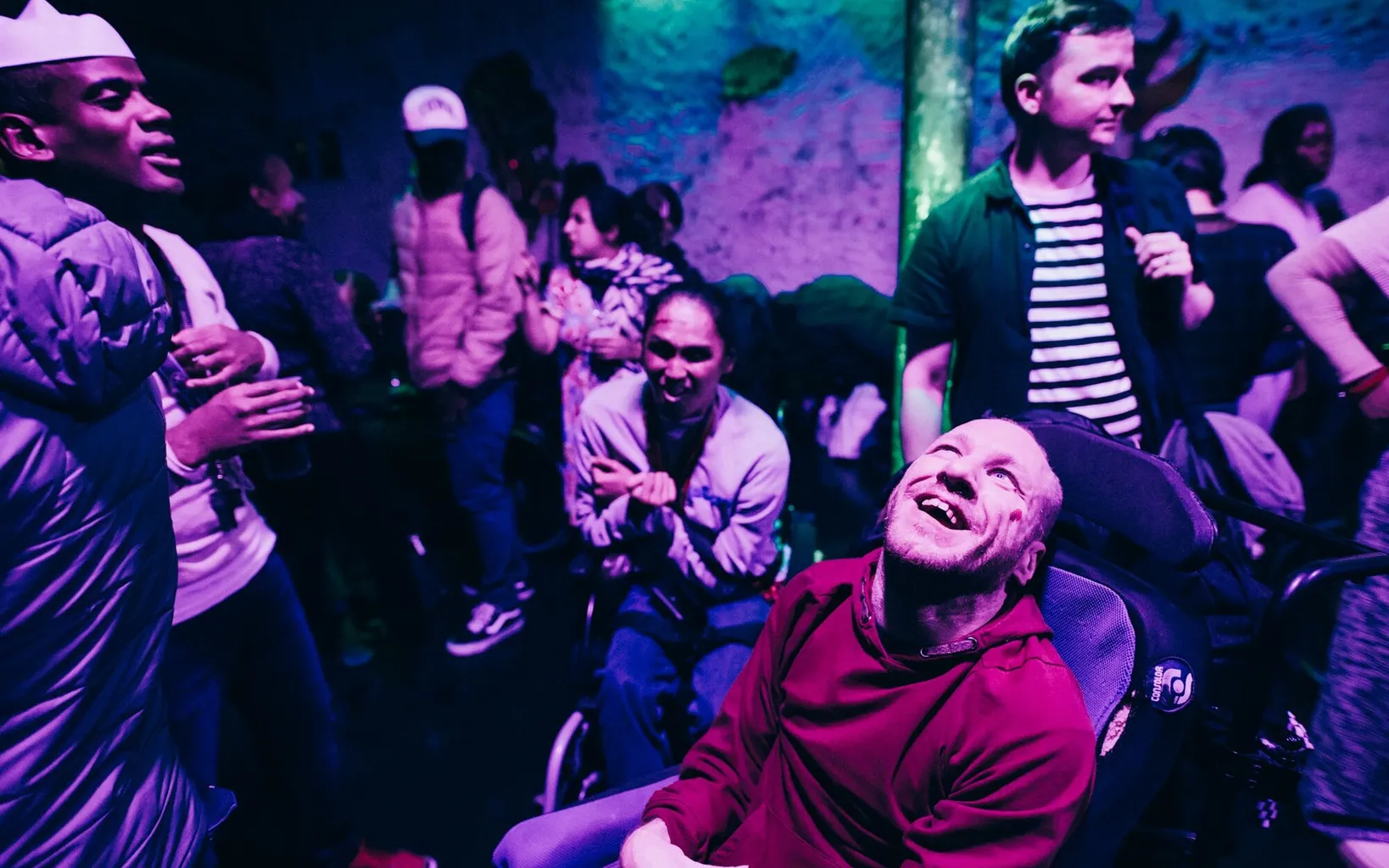 A group of people, some in wheelchairs, socialize and laugh together in a dimly lit club with purple lighting.
