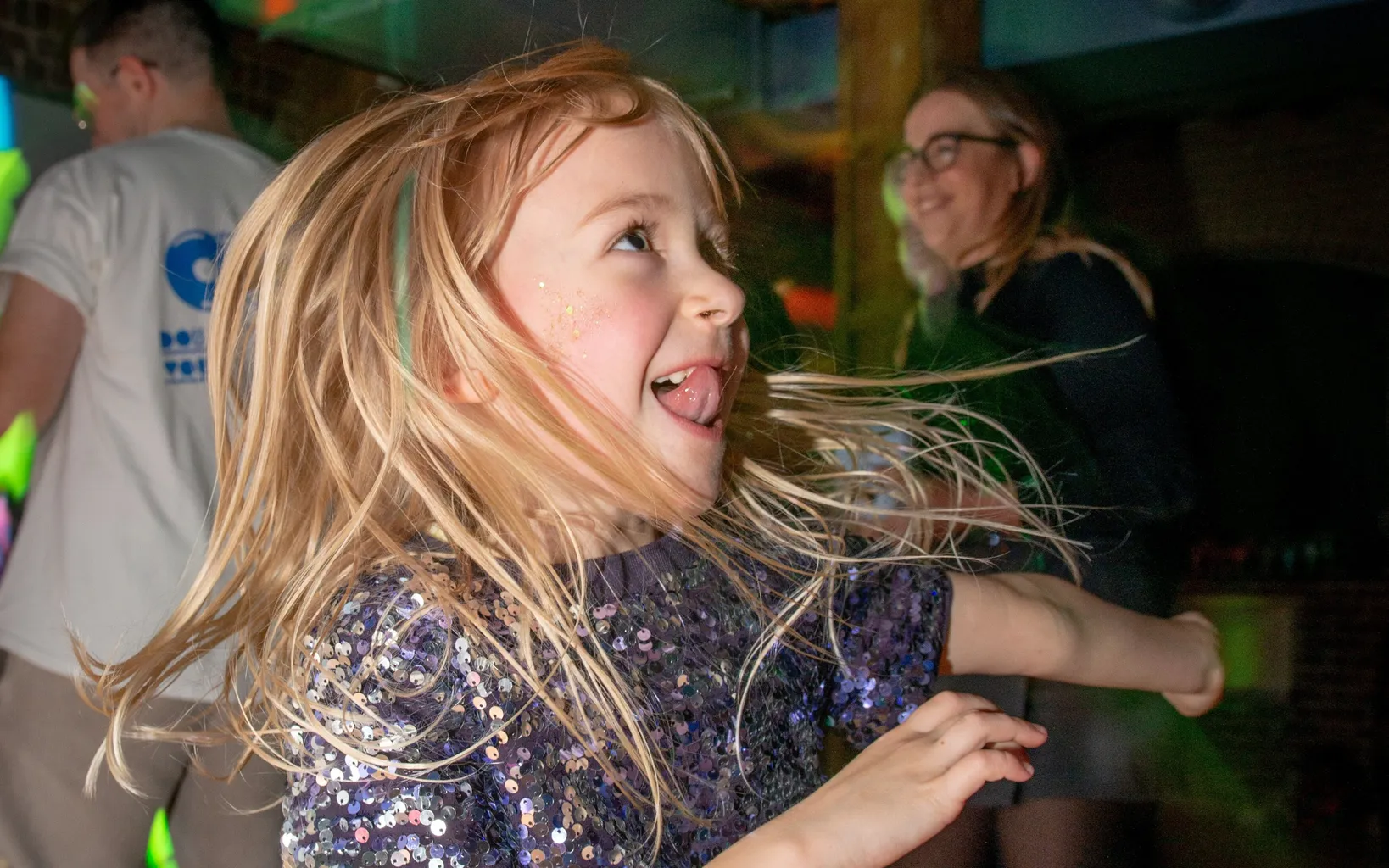 A young girl in a sparkly dress joyfully dances in a room with colorful lights. Two adults are in the background.