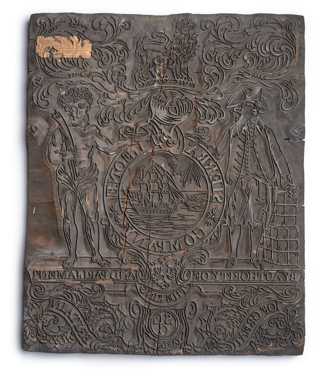 An antique wooden printing block featuring intricate carvings of two figures, text, and decorative elements with a damaged section at the top left corner.