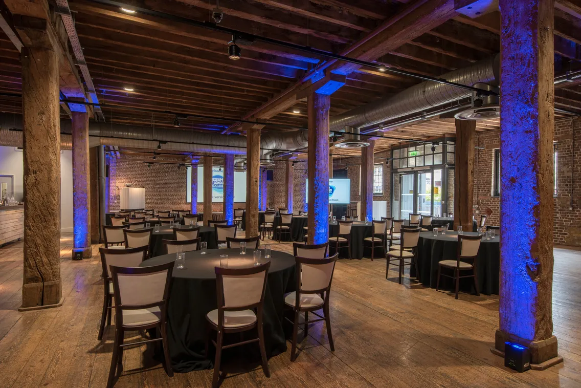 Spacious venue with wooden beams, round tables covered in black cloths, and chairs. Dim lighting and blue accent lights on walls create a warm atmosphere.
