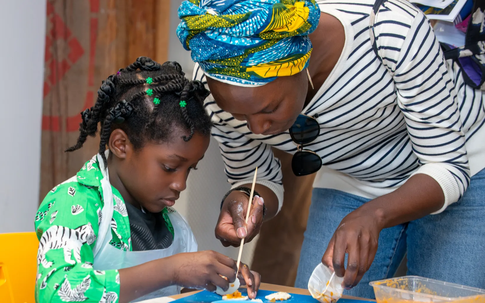 A child wearing a bright green patterned top and apron is seated at a table with dough and flour laid out in front of her. Over her, a woman is bent over, painting the dough with a pastry brush.
