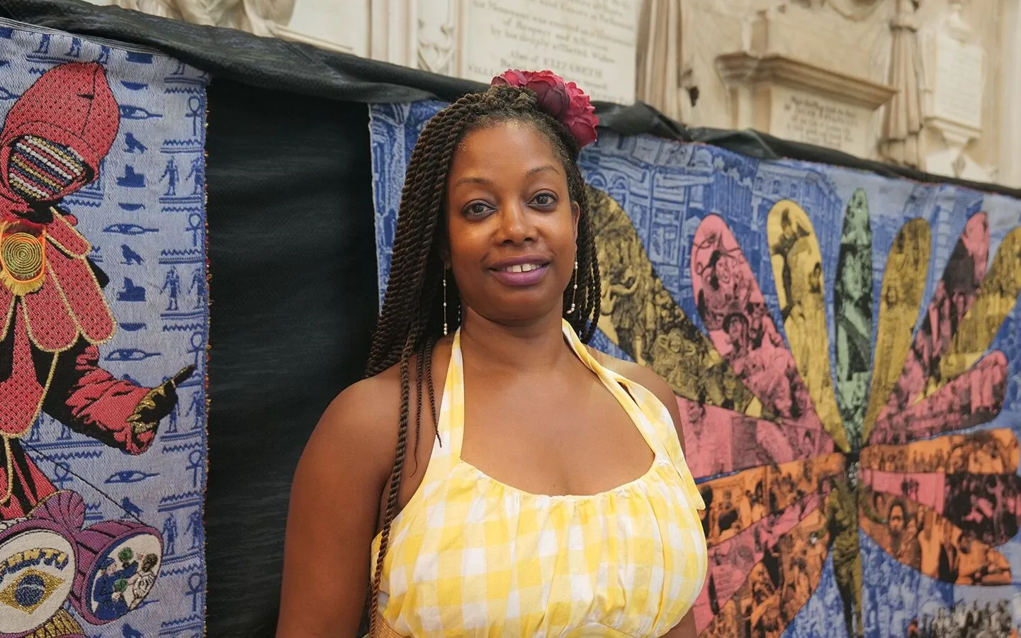 A woman in a yellow checkered dress stands in front of a colourful, tapestry artwork featuring intricate patterns and historical Caribbean imagery.