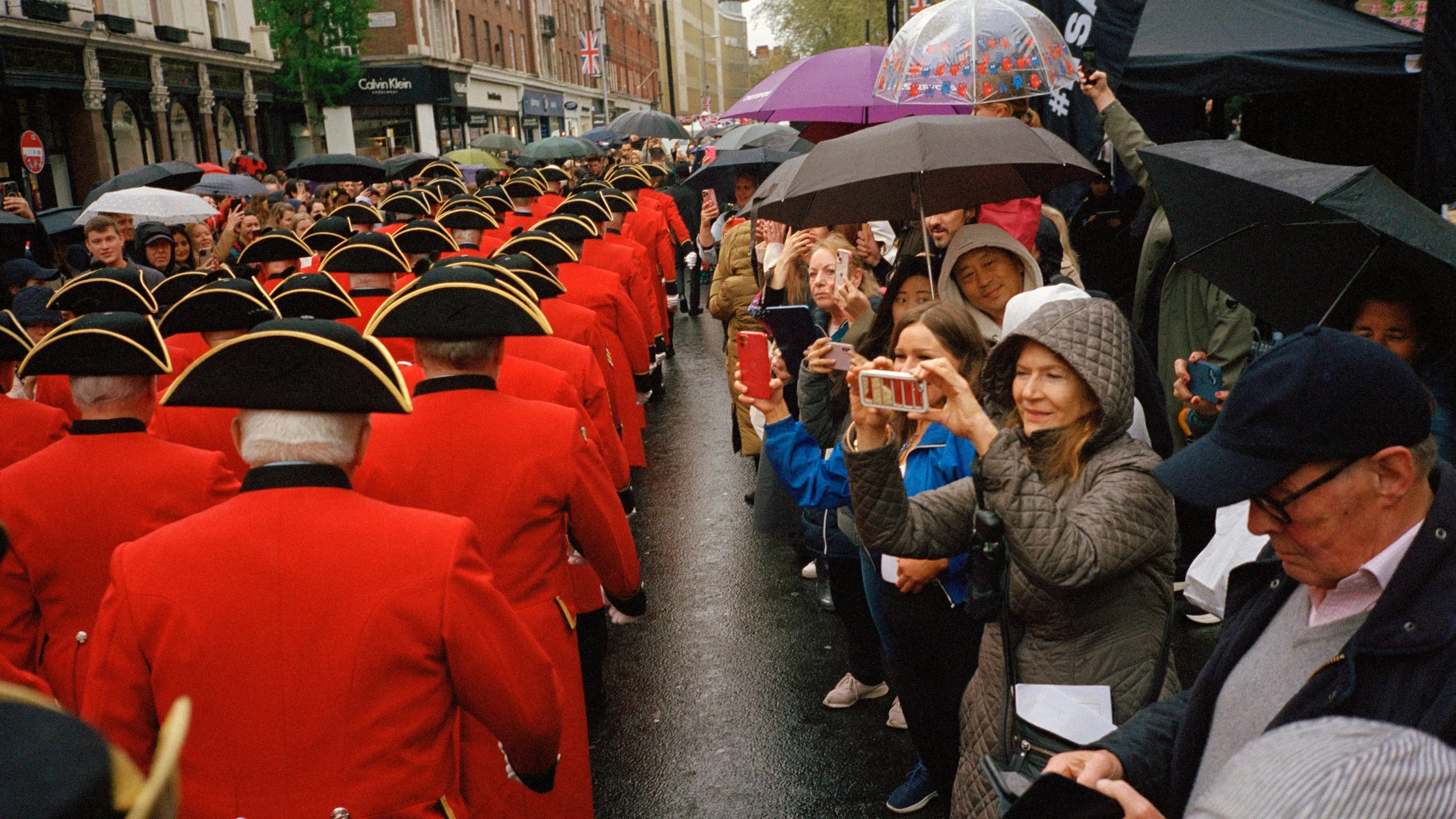 Digital photograph. Scanned from the negative. The King’s Coronation, Kings Road, Chelsea, UK. 
The Chelsea Pensioners march up Kings Road in formation, rain pouring, to serenade the King as the public spectate.