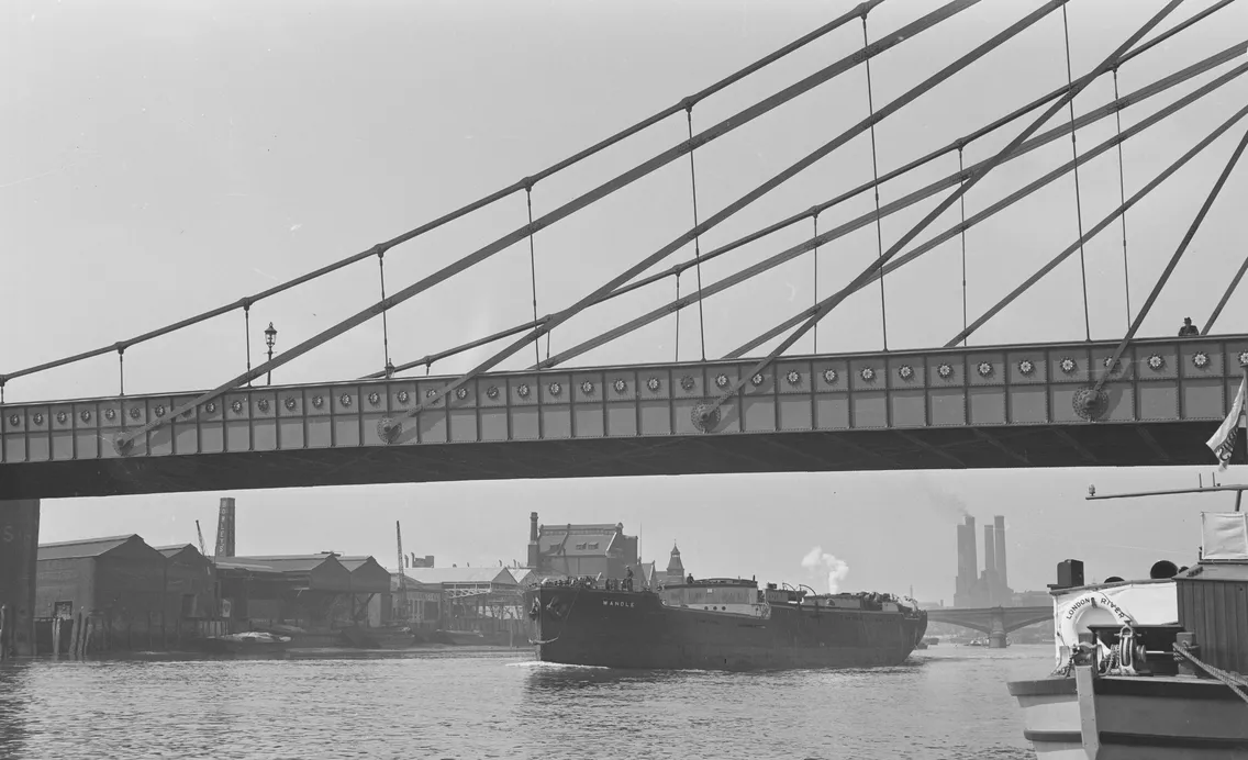 Black and white image of a cargo ship passing under a suspension bridge in an industrial harbor.