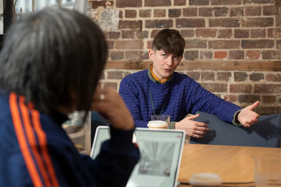 Two people sitting at a table, having a conversation. One gestures while speaking, and the other listens, partially visible from behind. A laptop and coffee cup are on the table.