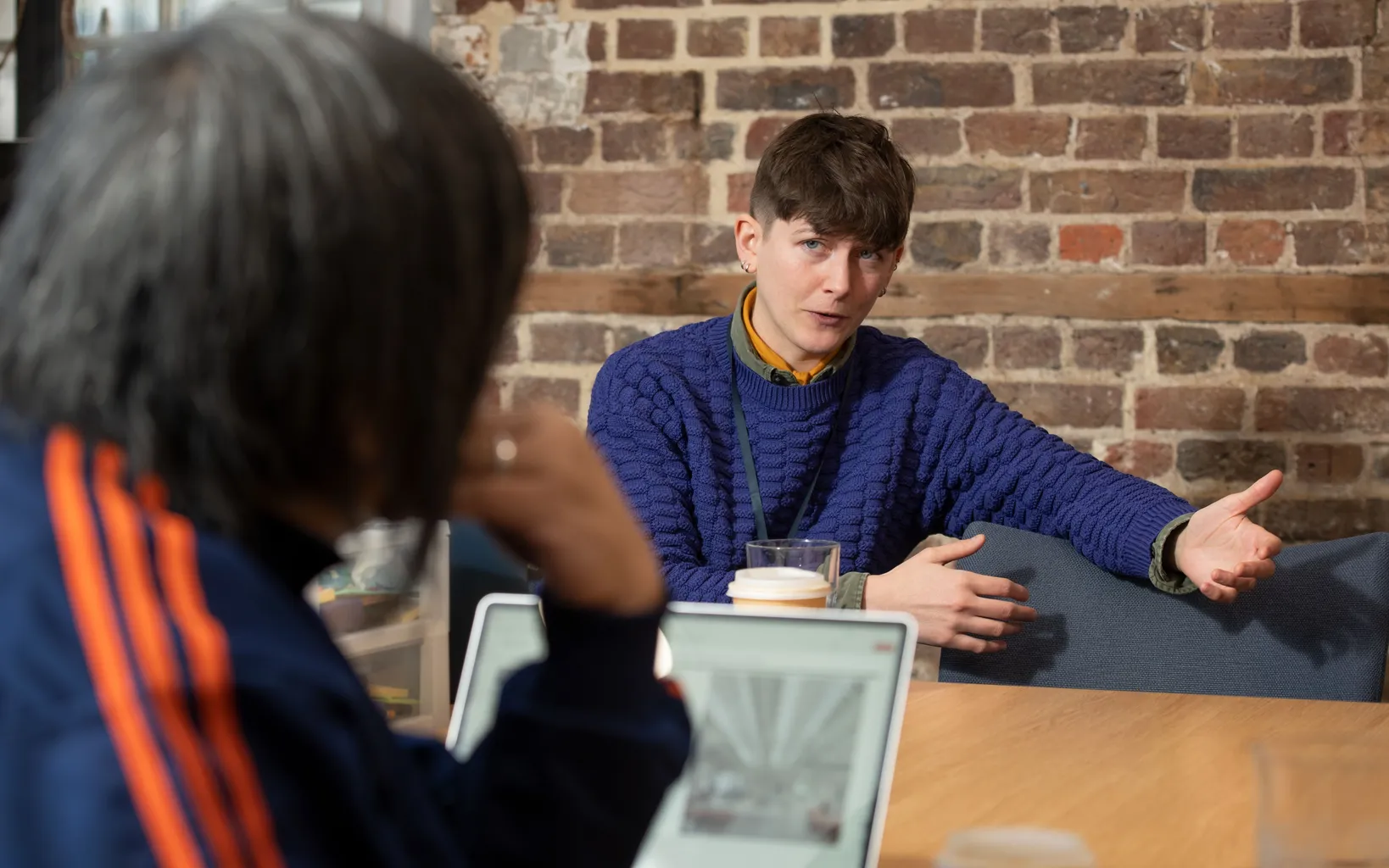 Two people sitting at a table, having a conversation. One gestures while speaking, and the other listens, partially visible from behind. A laptop and coffee cup are on the table.