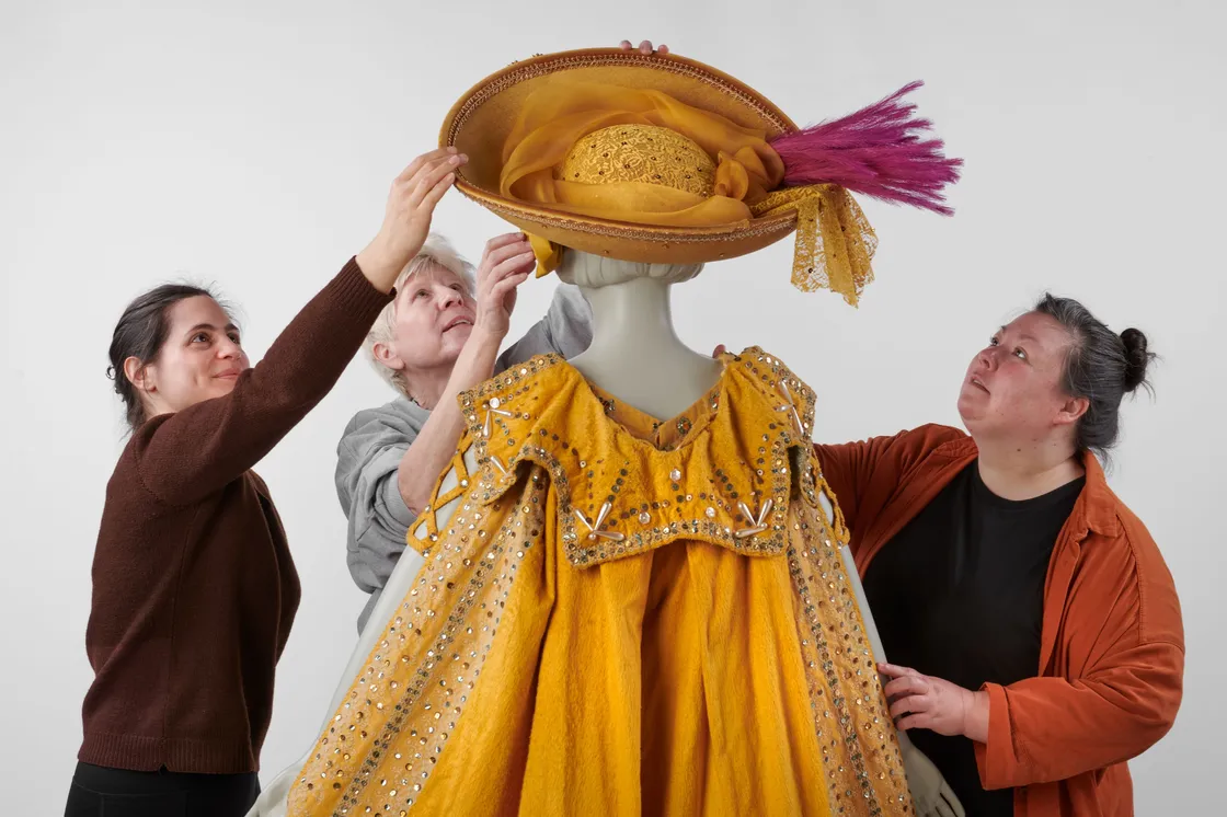 Three people adjust a large, elaborate yellow hat and matching ornate yellow costume on a mannequin against a plain background.