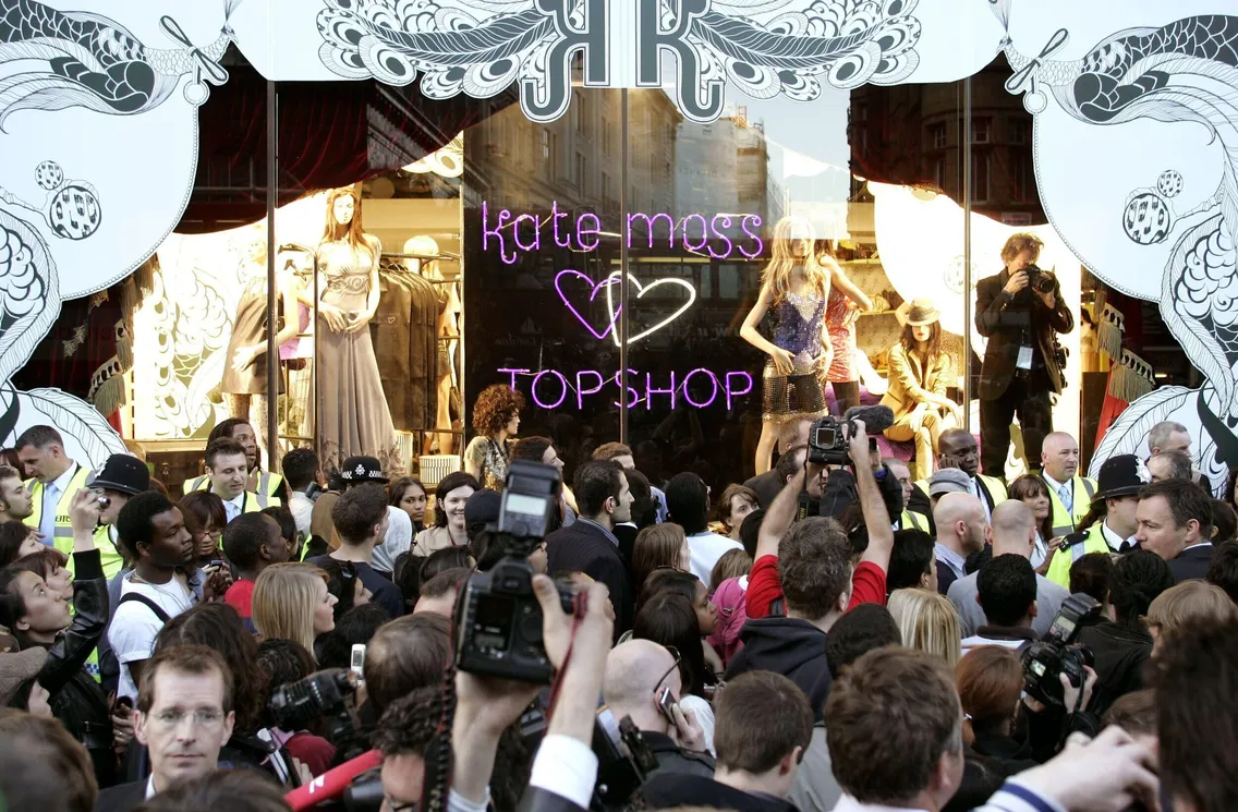 A large crowd and photographers wait outside a Topshop store window display with mannequins and a sign reading “Kate Moss ♥ TOPSHOP,” eager for the new collection.
