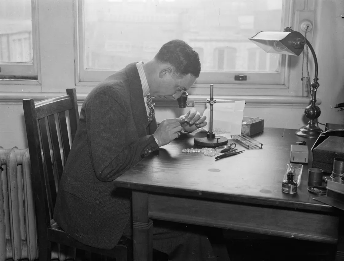 A man in a suit sits at a desk examining gemstones with tools and a magnifying device, in a well-lit room with large windows.