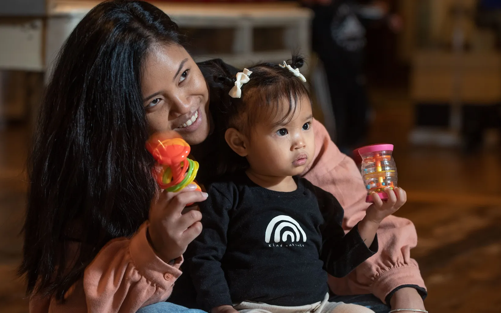 A woman with long, straight black hair sits on the ground with a toddler girl in her lap. The toddler wears a black, long-sleeved top with a white rainbow. Both hold multicoloured shakers in their hands.