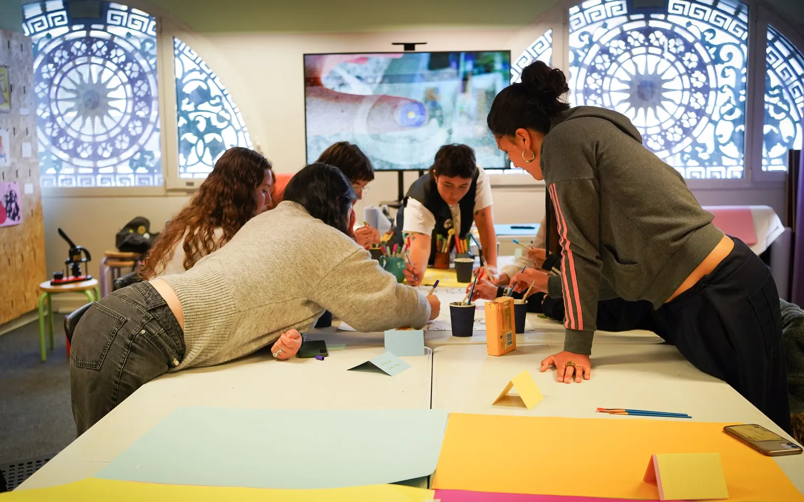 Five people gather around a table covered with colorful papers, markers, and sticky notes, engaging in a collaborative group activity in a well-lit room with decorative windows. Among them, ELENI leads the discussion while referencing resources from
