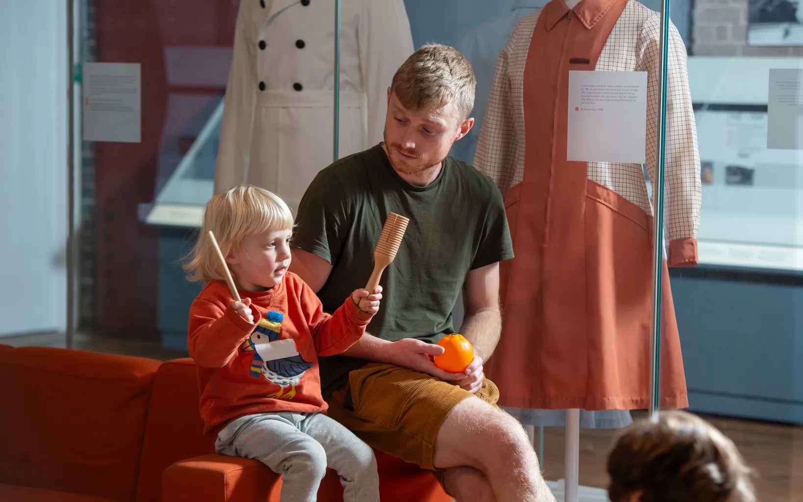 A young child and an adult sit on an orange bench; the child holds percussion instruments while the adult watches, with mannequins wearing uniforms displayed in the background.