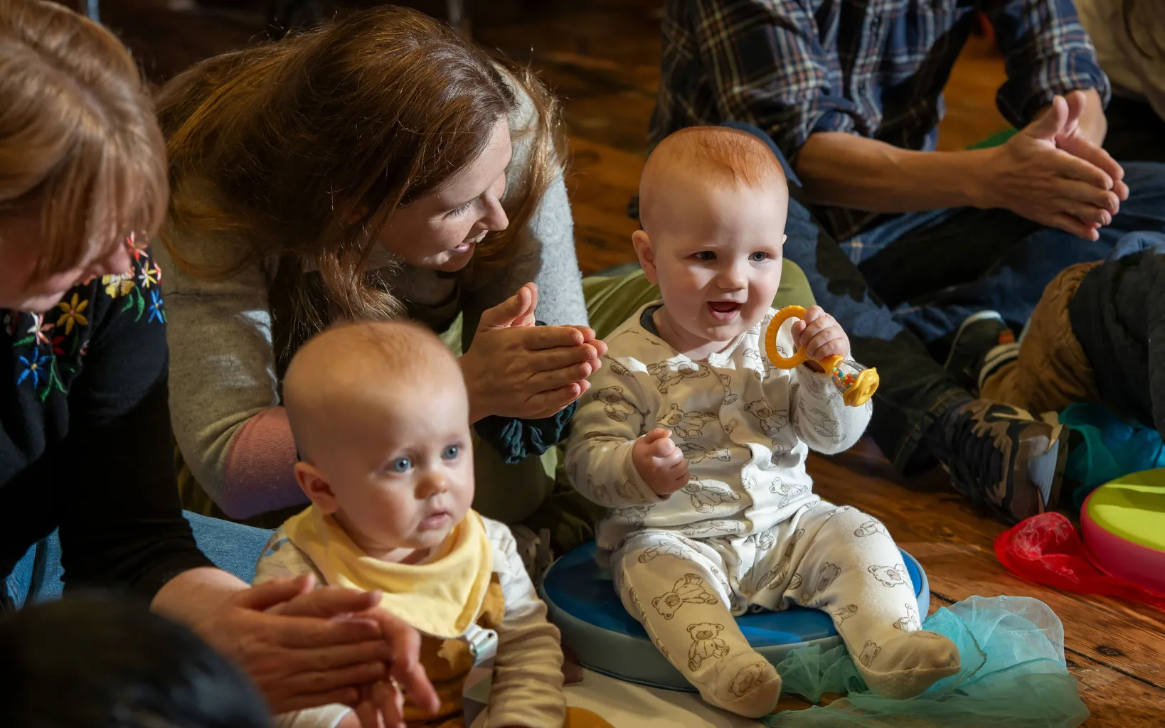 Two babies sit on the floor among adults; one baby holds a teething toy and smiles, while the other looks ahead. Adults are clapping and sitting close to the children.