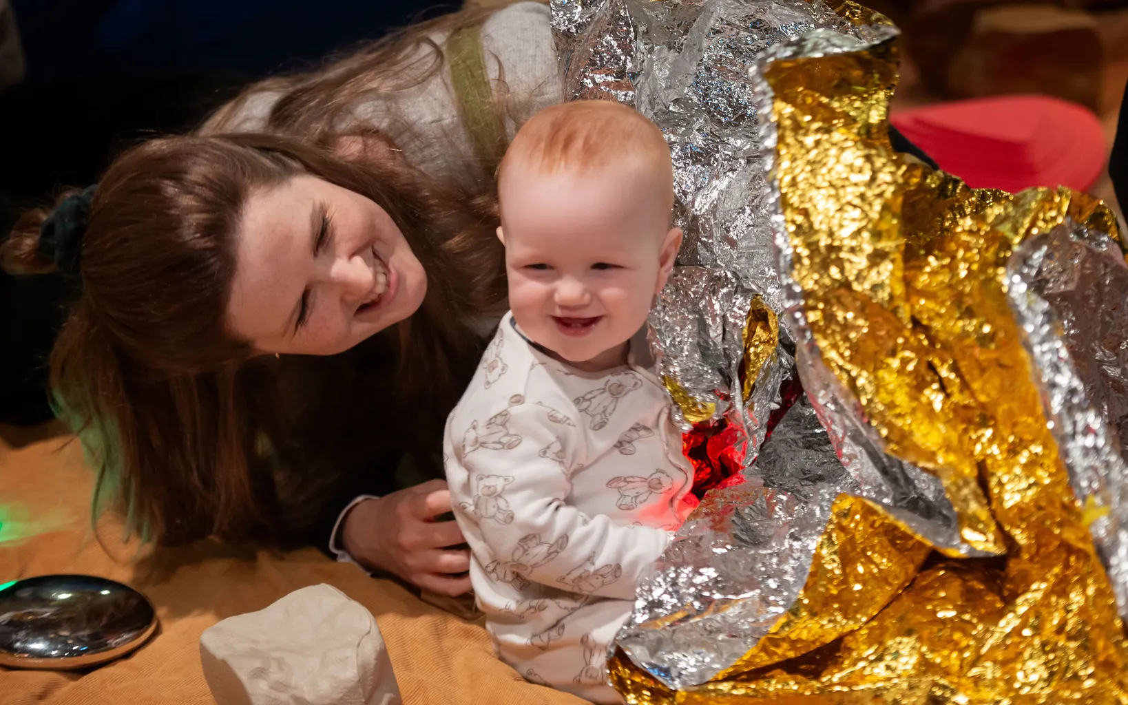 An adult and a smiling baby sit on the floor with shiny silver and gold foil blankets, lit by a small red light.