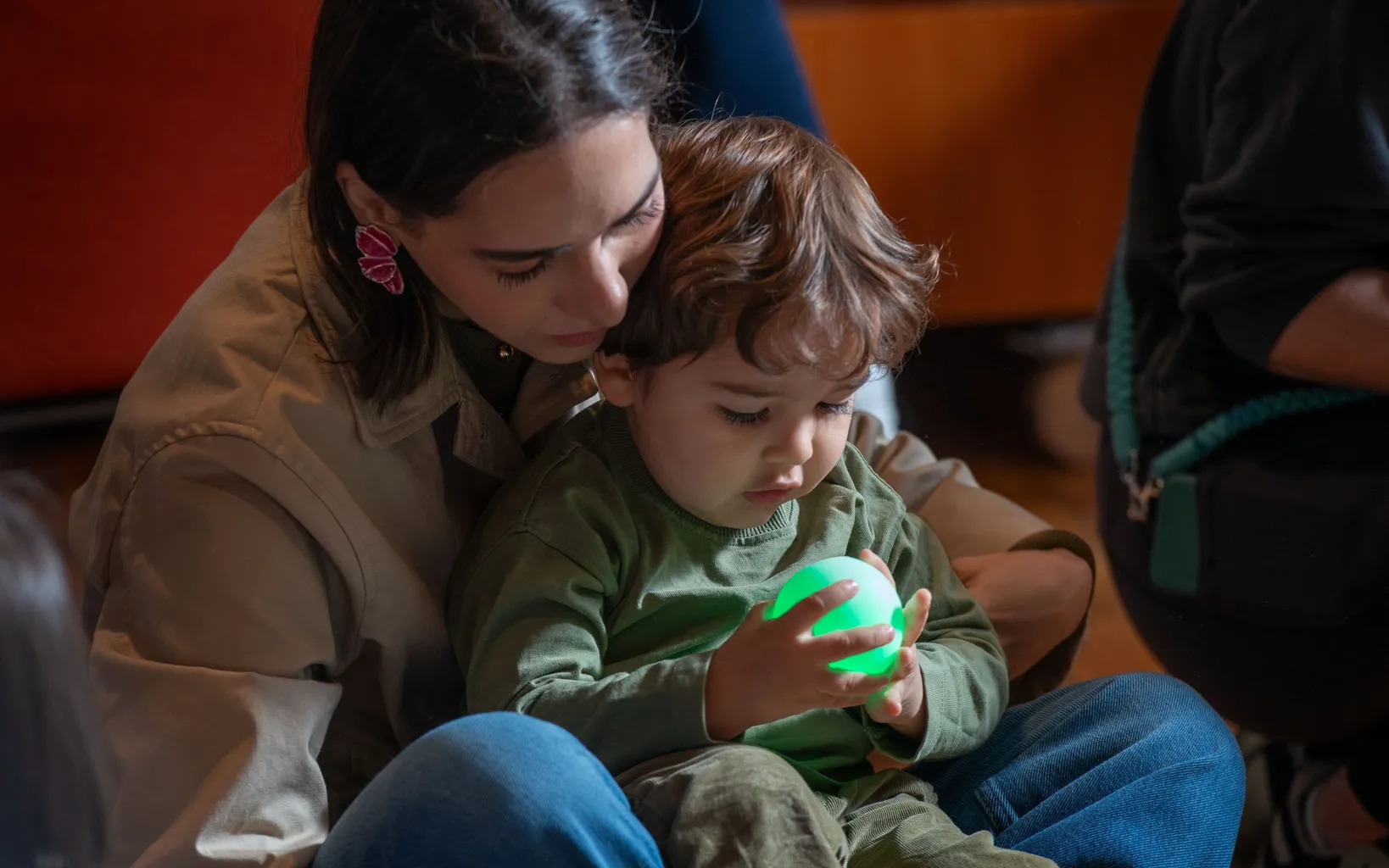 An adult sits with a young child on their lap. The child is holding and looking at a glowing green ball.