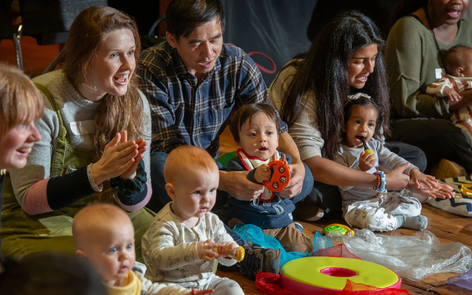 Adults and children interact energetically in a room with musical instruments and sheet music, while a violinist plays in the foreground.
