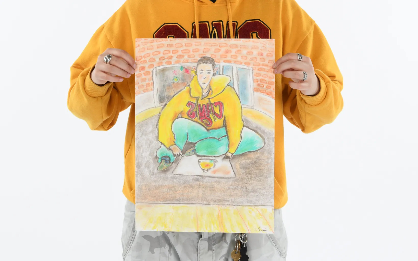 A person in a yellow sweatshirt holds a colorful drawing of someone wearing a similar sweatshirt and sitting on the floor with art supplies.