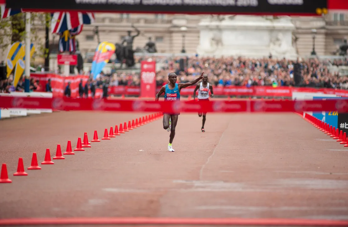 Two people about the cross a red banner as part of a marathon race, with one person waving at a crowd