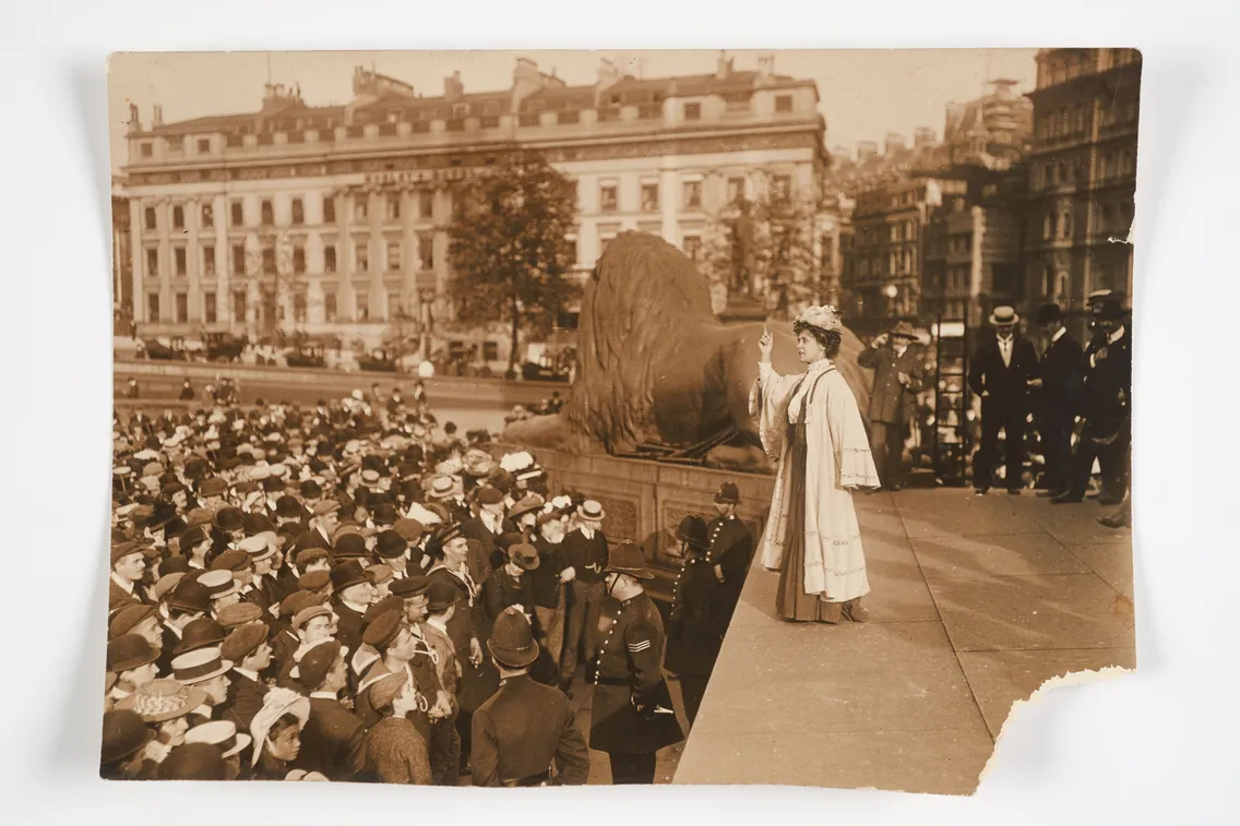 Woman in white dress addressing a large crowd at a public square near a lion statue, in a historical sepia-toned photograph.