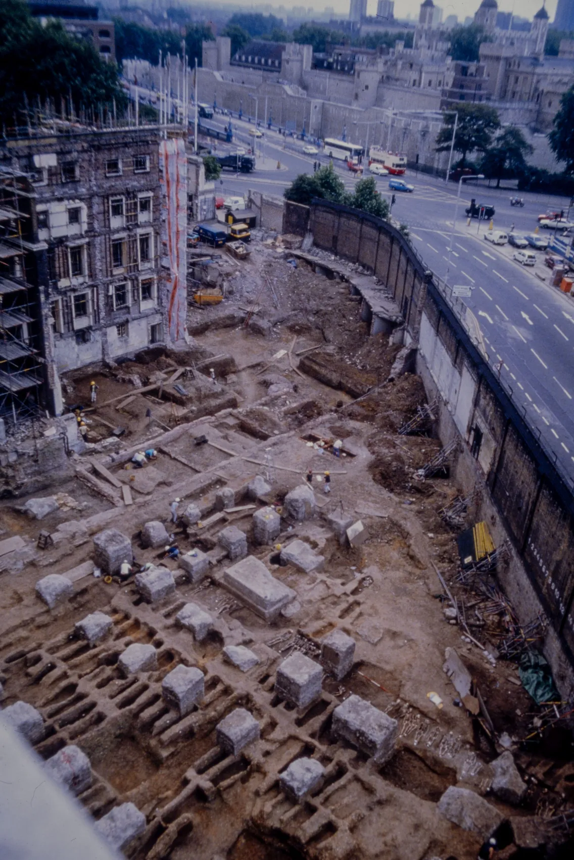 Archaeological excavation site beside a busy road, with exposed stone foundations and a partially scaffolded building nearby.