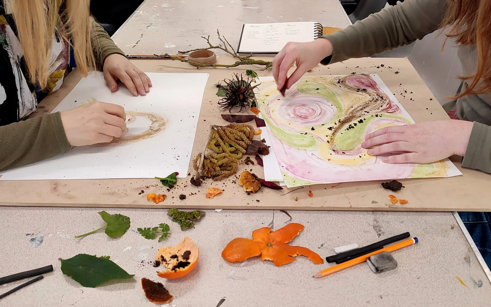 Two people sat opposite each other at a table while they draw, with earthy materials around them including leaves and orange peel