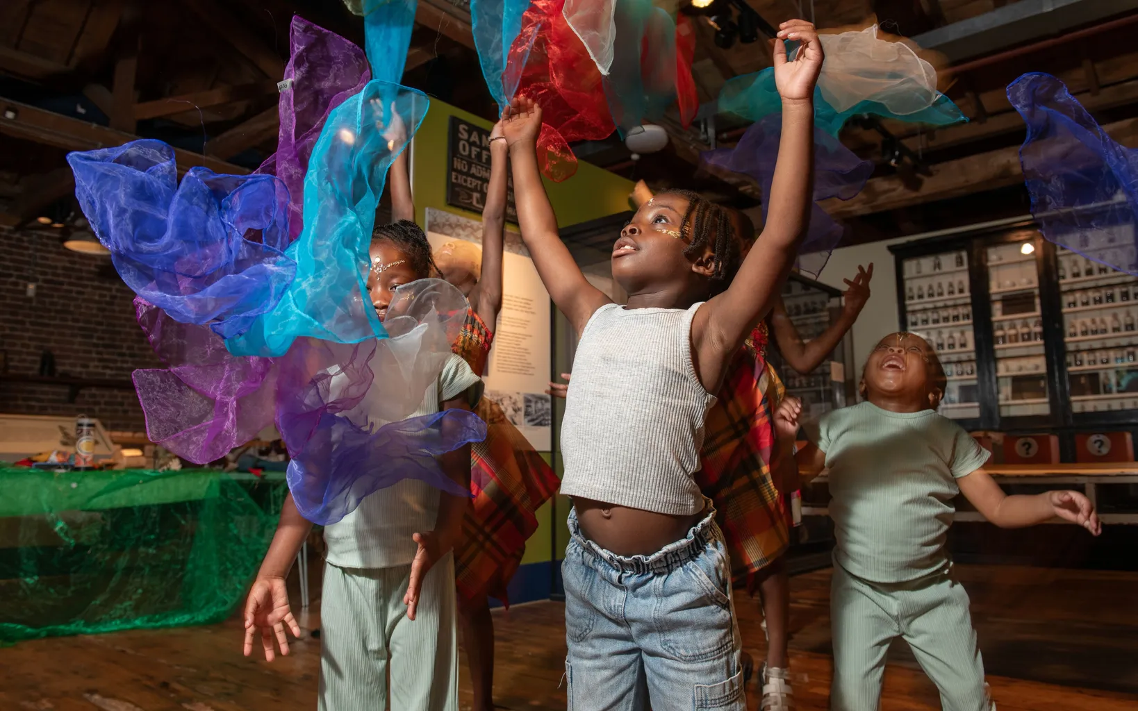 A woman in colorful clothing speaks animatedly to a group of young children seated on the floor in a classroom or community space.
