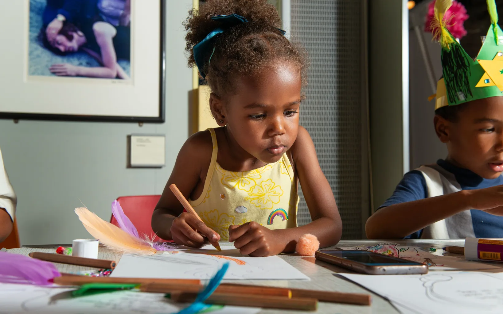 A young girl sits at a table drawing with colored pencils, surrounded by art supplies, next to another child wearing a paper crown, as a Caribbean family festival takes place around them.