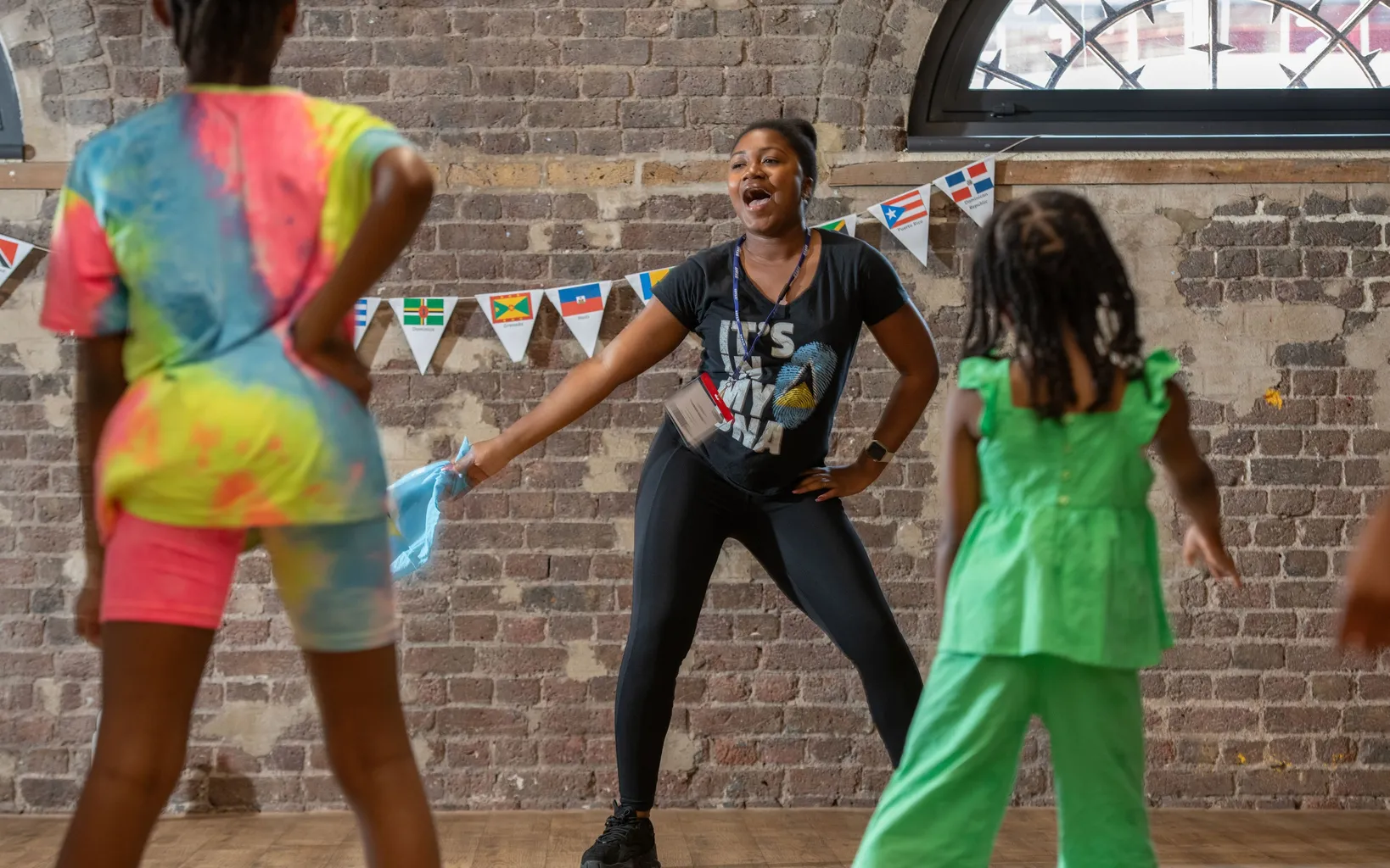 An instructor leads two children in a dance or exercise activity indoors, celebrating culture with international flag bunting hanging on a brick wall in the background.