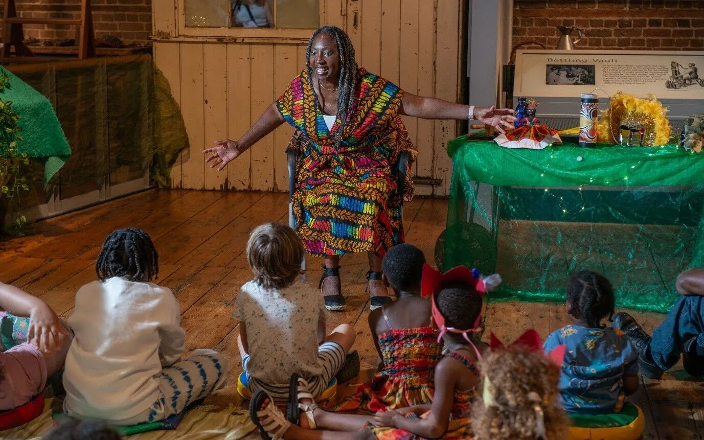 A woman in colorful clothing speaks animatedly to a group of young children seated on the floor in a classroom or community space.