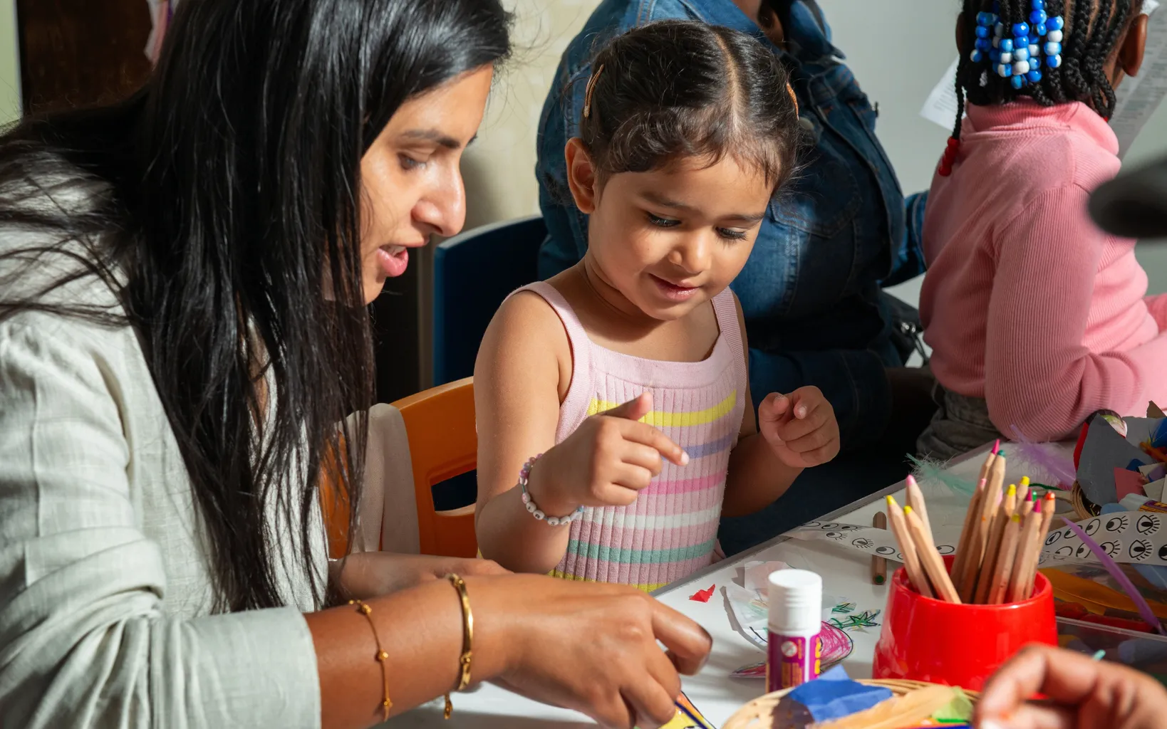 An adult and a young girl work on an arts and crafts. project at a table with colored pencils and supplies, surrounded by other children, sharing family moments and celebrating creativity as part of a vibrant cultural festival.