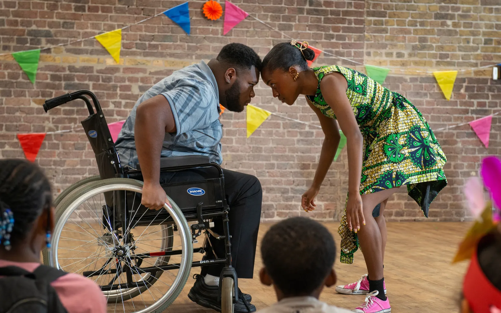 A man in a wheelchair and a woman stand head to head in front of a brick wall decorated with colorful pennant banners, celebrating Caribbean culture as children watch.