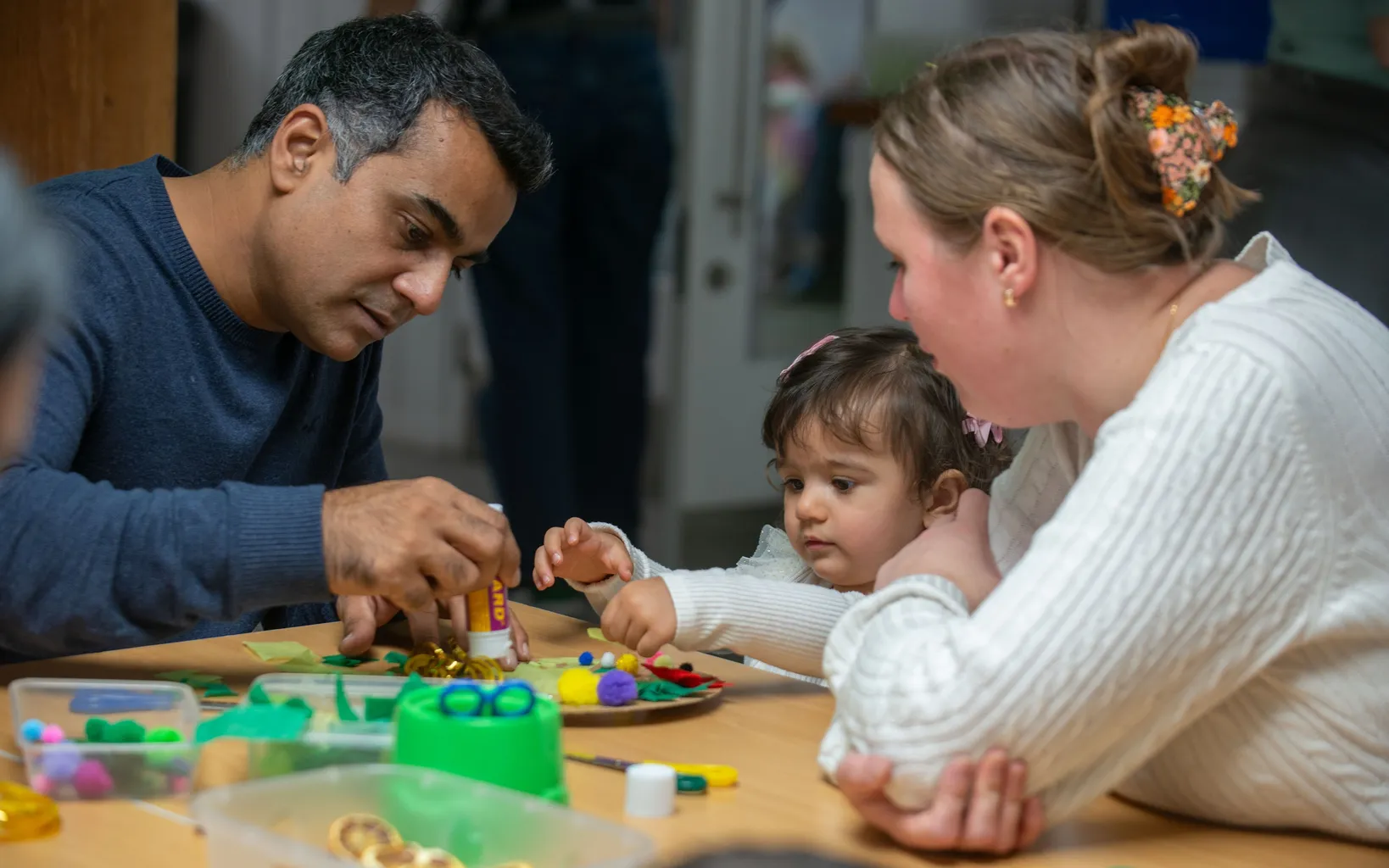 Two adults and a child engage in a craft activity at a table with various materials.